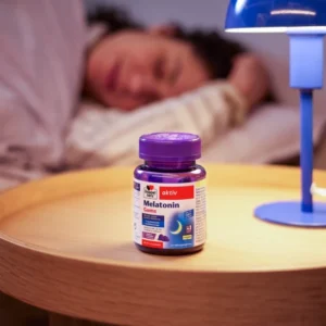 Лилава опаковка „Doppelherz aktiv Melatonin – Gums/Желирани таблетки“ върху дървено нощно шкафче до синя лампа; на заден план човек спи в легло. Фокус върху продукта за по-лесно заспиване.